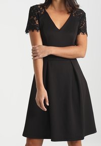 Abito nero con scollo a V e maniche corte in pizzo. La gonna si allarga dalla vita, caratterizzata da una texture liscia e un design semplice.