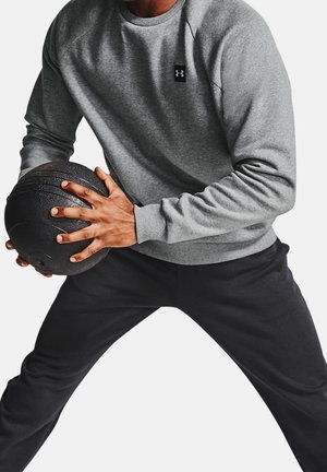 Personne portant un sweat-shirt gris Under Armour et un pantalon noir tenant un ballon médicinal noir à texture des deux mains en position athlétique.