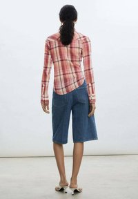 Red and pink plaid long-sleeve shirt with fitted shoulders, paired with loose denim bermuda shorts. Model wears white heeled mules.
