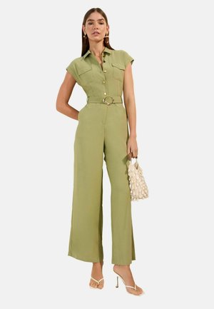 Nő olíva zöld, öves overált visel rövid ujjakkal, krém színű kagyló alakú kézitáskát tart, és fehér magas sarkú szandált visel.