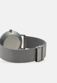 Metallklocka med ett runt grått boett och ett mesharmband. Baksidan är slät och metallisk. Armbandet har en texturerad, vävd design.