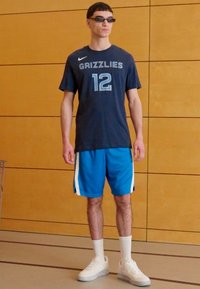 Marineblå sports t-shirt med "Grizzlies" og nummer "12", sammen med blå shorts med hvide striber. Bærer sorte solbriller og hvide sneakers.