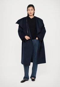 Manteau en laine bleu marine avec de larges revers, associé à un pull noir texturé et un denim bleu. Le modèle porte des ballerines noires et se tient contre un fond clair.