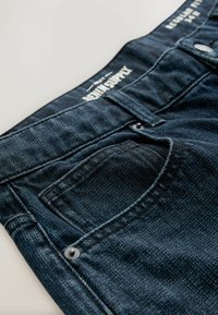 Tumed denim teksased, millel on esikott, hõbedane riistvara ja õmmeldud vöökoht brändingu vahemikus. Tekstuuri on sile ja klassikaline disain.