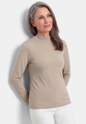 Femme aux longs cheveux gris portant un pull à col roulé beige à manches longues et un pantalon blanc, debout devant un fond gris clair.