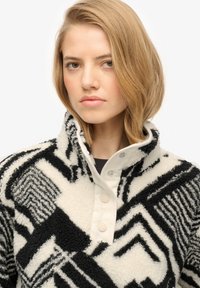 Jonge vrouw met kort blond haar die een zwart-witte fleece trui met een patroon draagt, voorzien van drukknopen op de kraag.