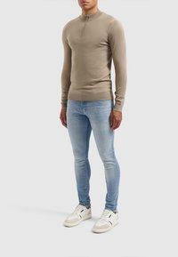 Ljusbeige långärmad tröja med zipkrage, i kombination med ljusblå snäva jeans och vita sneakers, som visar upp en figurnära design och slät textur.