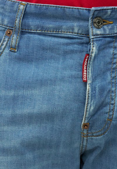 Κοντινή λήψη της μπροστινής τσέπης μπλε τζιν με πορτοκαλί ραφές, μεταλλικά κουμπιά και κόκκινη ετικέτα της μάρκας DSQUARED2.