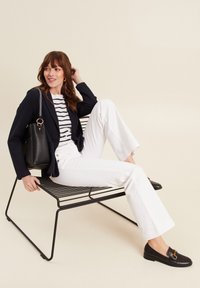 Blazer nero su una camicia a righe, pantaloni bianchi a palazzo, mocassini neri e una borsa nera dal design strutturato, seduto su una sedia in metallo contro uno sfondo semplice.
