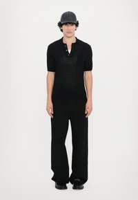 Schwarzes kurzärmliges Polohemd mit einem strukturierten Muster und einer Knopfleiste mit drei Knöpfen, kombiniert mit weit geschnittenen schwarzen Hosen und glänzenden Schuhen.