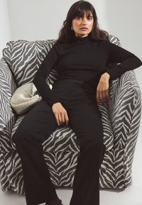 Melna pieguļoša garām piedurknēm blūze un melnas platās bikses, sēžot uz rakstainas pelēkas un baltas zebra raksta krēsla. Strukturēta krēmkrāsas rokassoma.