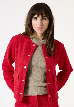 Veste rouge texturée avec des boutons-pression dorés et des poches, superposée sur un haut fuzzy beige clair. Tissu lisse avec une apparence confortable.