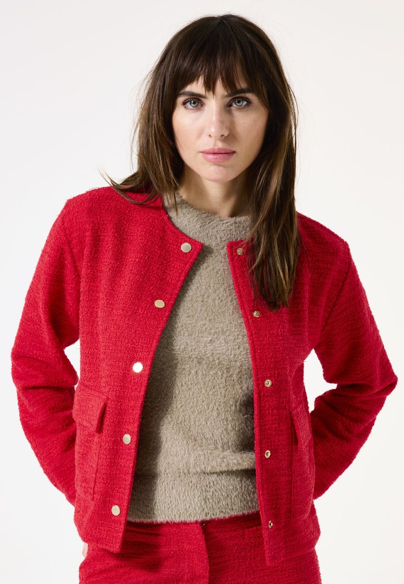 Veste rouge texturée avec des boutons-pression dorés et des poches, superposée sur un haut fuzzy beige clair. Tissu lisse avec une apparence confortable.