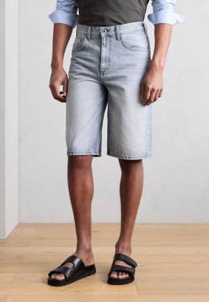 Άνδρας που φοράει ανοιχτό μπλε τζιν σορτς, μαύρα σανδάλια slide και ένα πουκάμισο με μακριά μανίκια γυρισμένα, στέκεται σε ξύλινο πάτωμα.