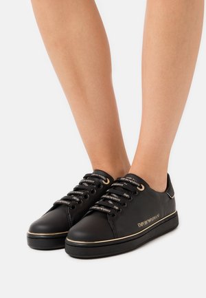 Sneakers basse in pelle nera con logo "Emporio Armani" in oro, lacci coordinati con il marchio e profili dorati sulla suola indossate ai piedi.
