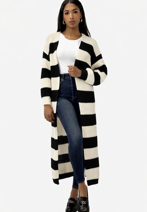 Mujer de pie vistiendo un cárdigan largo a rayas negras y blancas, camiseta blanca, vaqueros ajustados oscuros y mocasines negros con detalle dorado.