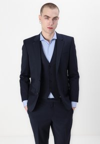 Marineblauw driedelig pak, getailleerde pasvorm, met een bijpassend vest. Lichtblauw overhemd met kraag zichtbaar eronder. Beschikt over klassieke knoopdetails.