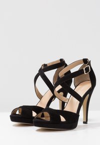 Sandales à talons hauts en daim noir avec des straps croisées, bout ouvert et une semelle intérieure lisse beige avec un talon aiguille fin.