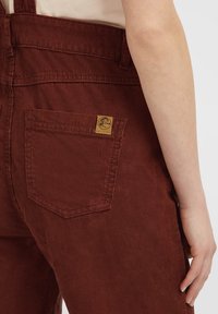 Peto de pana marrón que presenta un bolsillo trasero con una etiqueta de marca cuadrada. La tela tiene una textura acanalada visible y un diseño de corte relajado.