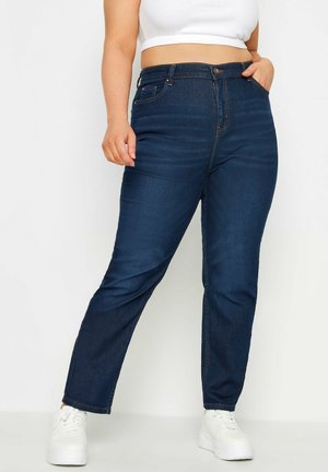 Kvinde iført mørkeblå højttaljedejeans, hvid cropped top og hvide plateausneakers, står med den ene hånd i lommen.