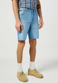 Pantaloni corti in denim di colore azzurro chiaro, con un taglio classico, design a cinque tasche e una texture sbiadita. Indossati con scarpe in suede beige e calze bianche.