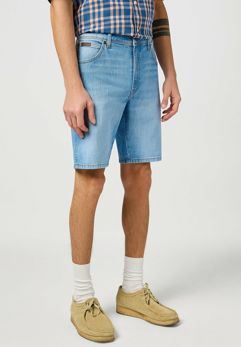 Pantaloni corti in denim di colore azzurro chiaro, con un taglio classico, design a cinque tasche e una texture sbiadita. Indossati con scarpe in suede beige e calze bianche.