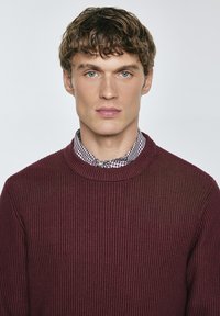 Bordeauxfarbener, gerippter Strickpullover mit rundem Halsausschnitt, über einem schwarz-weißen karierten Hemd getragen, mit mittelschwerer Stoffstruktur.