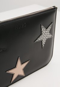 Trousse noire à fermeture éclair avec des étoiles décoratives cousues, une beige et une à motif avec texte répété, et nom de marque embossé "HUGO."