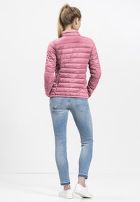 Lichtroze pufferjack met een hoge kraag en een gestikt patroon, gecombineerd met lichtblauwe skinny jeans en witte sneakers.