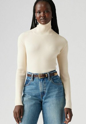 Ribbed turtleneck sweater in light beige, paired with high-waisted blue jeans and a brown belt featuring a gold buckle.