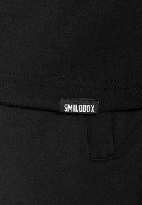 Tejido elástico negro que presenta una etiqueta visible que dice "SMILODOX." La prenda tiene una textura suave y un diseño simple sin patrones distintivos.