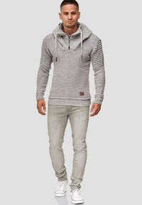 Grijze hoodie met een ribbeltextuur en vooraan een rits. Voorzien van trekkoorden, zijdelingse details en een logopatch. Draagt met lichtgrijze jeans en witte sneakers.