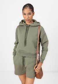 Olivgrünes Hoodie- und Shorts-Set aus weichem Stoff; mit einer Kordelzugkapuze, Kängurutasche und gerippten Bündchen. Braune Umhängetasche.