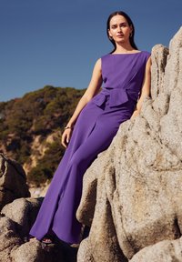 Lila overál, strukturált felső résszel, széles szárú szabással és masni díszítéssel a deréknál. Simára szabott anyag textúrált sziklás háttér előtt.