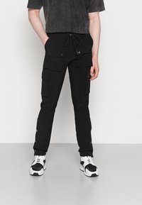 Person som bär svarta cargobyxor med stora knappliknande fickor och svart-vita sneakers, stående mot en enkel ljus bakgrund.