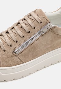 Baskets en daim beige avec une semelle texturée blanche, dotées de lacets plats et d'un détail de fermeture éclair sur le côté avec un tirette métallique.