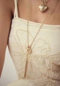 Woman wearing a cream dress with sequin floral patterns and two gold necklaces, one with a heart pendant and one with chain tassels.