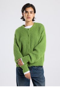 Green knitted cardigan with a ribbed texture, seven buttons, and long sleeves. White cuffs peeking from under the sleeves. Casual fit.