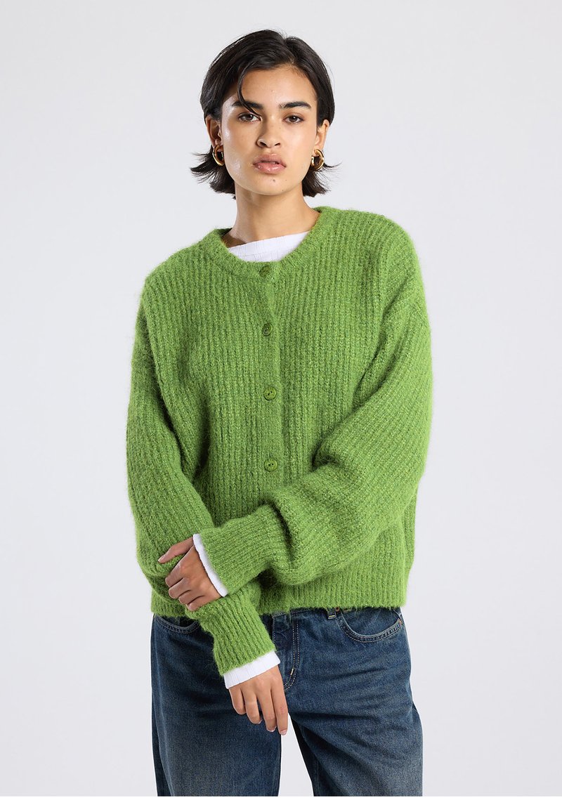 Green knitted cardigan with a ribbed texture, seven buttons, and long sleeves. White cuffs peeking from under the sleeves. Casual fit.