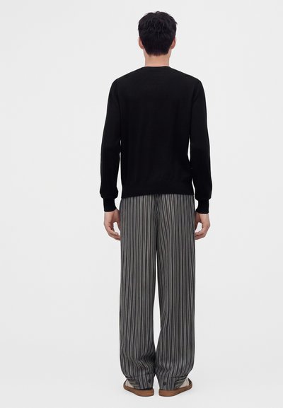 Homme debout de dos, portant un haut noir à manches longues, un pantalon large à rayures verticales gris et blanc, et des sandales sur un fond uni.