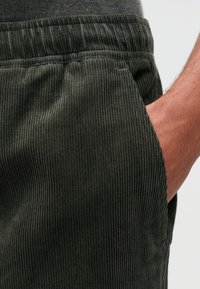 Groene corduroy broek met een textuur op het oppervlak, voorzien van een elastische tailleband met trekkoord en een zijzak, vastgelegd met een hand die op de stof rust.