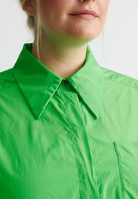 Heldengroen, lichtgewicht shirt met een puntige kraag, knoopsluiting en één borstzak. Gladde textuur met een subtiele glans.