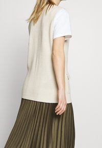 Beige stickad väst med ribbad textur, med en sidficka, bärs över en vit T-shirt och en mörkgrön plisserad kjol.