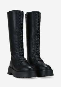 Botas de charol negras hasta la rodilla con suela gruesa, que presentan un diseño de cordones en la parte delantera y una cremallera lateral para facilitar su uso.