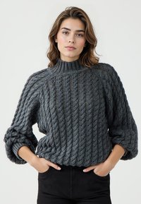 Donna che indossa un maglione a trecce grigio scuro con maniche arrotolate e pantaloni neri, in piedi con le mani nelle tasche.