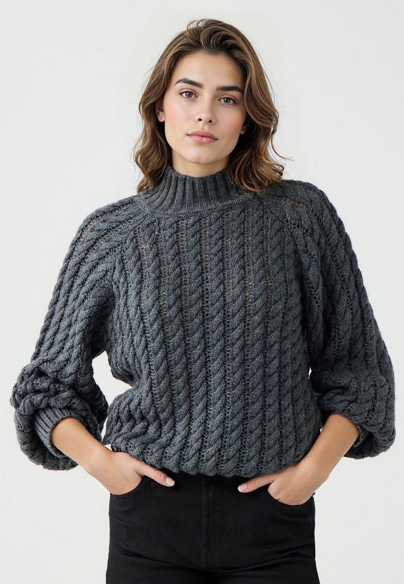 Donna che indossa un maglione a trecce grigio scuro con maniche arrotolate e pantaloni neri, in piedi con le mani nelle tasche.