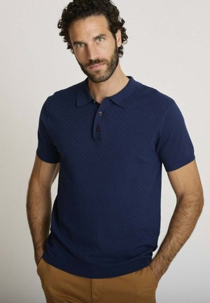 Homme aux cheveux bouclés foncés et à la barbe, portant un polo bleu marine à manches courtes et un pantalon marron clair, debout les mains dans les poches.