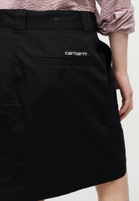 Άτομο που φοράει μαύρη φούστα Carhartt με πίσω τσέπη και κεντημένο λογότυπο, συνδυασμένη με ροζ πουκάμισο με υφή και μανίκια διπλωμένα προς τα πάνω.