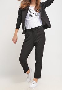 Sort zip-up jakke over en hvid grafisk t-shirt, parret med skræddersyede sorte bukser og hvide sneakers. Glat stof, minimalistisk design.