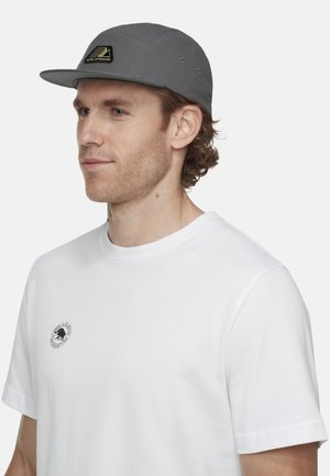 Man met lichtbruin krullend haar, een grijze baseballpet met logo en een wit T-shirt met een klein zwart rond logo op de borst.